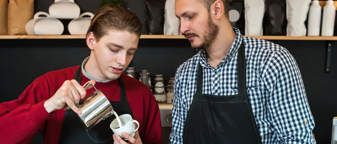 Professional barista teaching young man how to safely make cappuchino